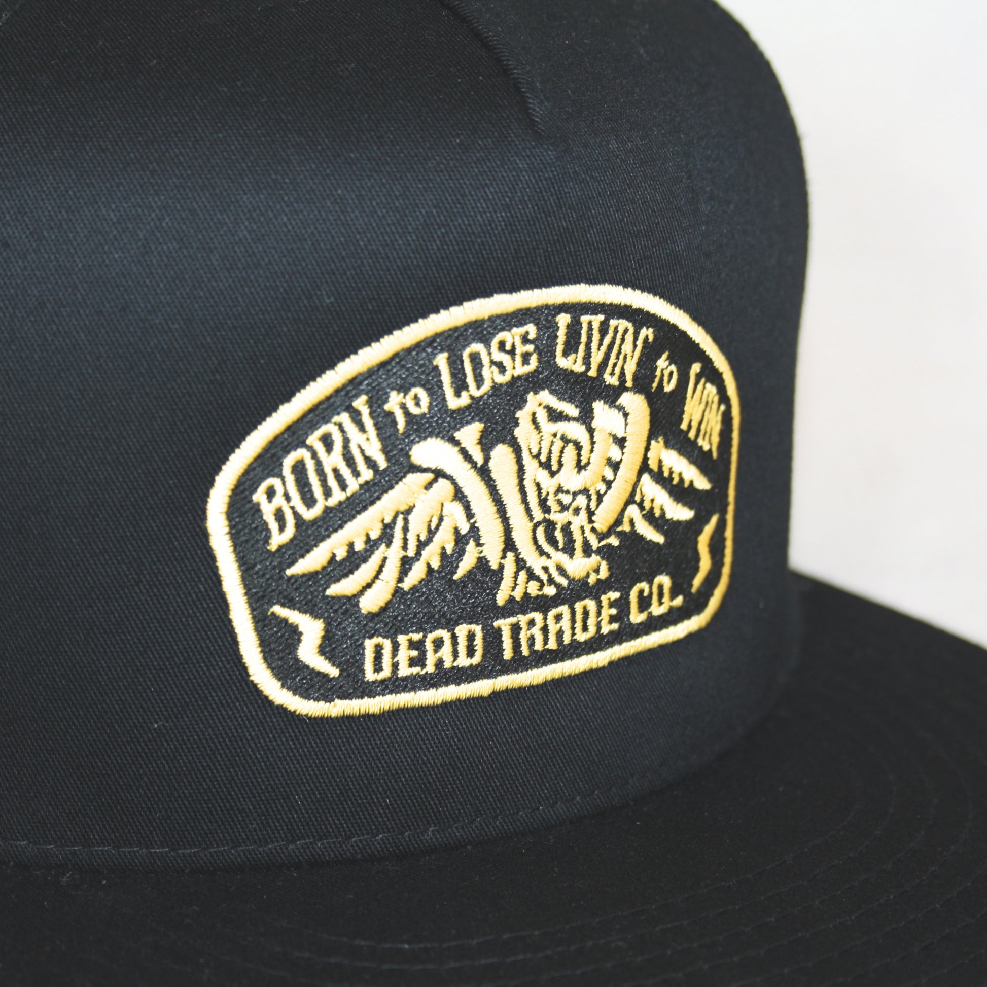 Black snapback with 'Born to Lose' gold embroidery on the front panel. High-profile, flat brim trucker cap with breathable mesh. Made of 65% polyester, 35% cotton. Structured 5-panel, 4” crown. Finished in Alberta, Canada.