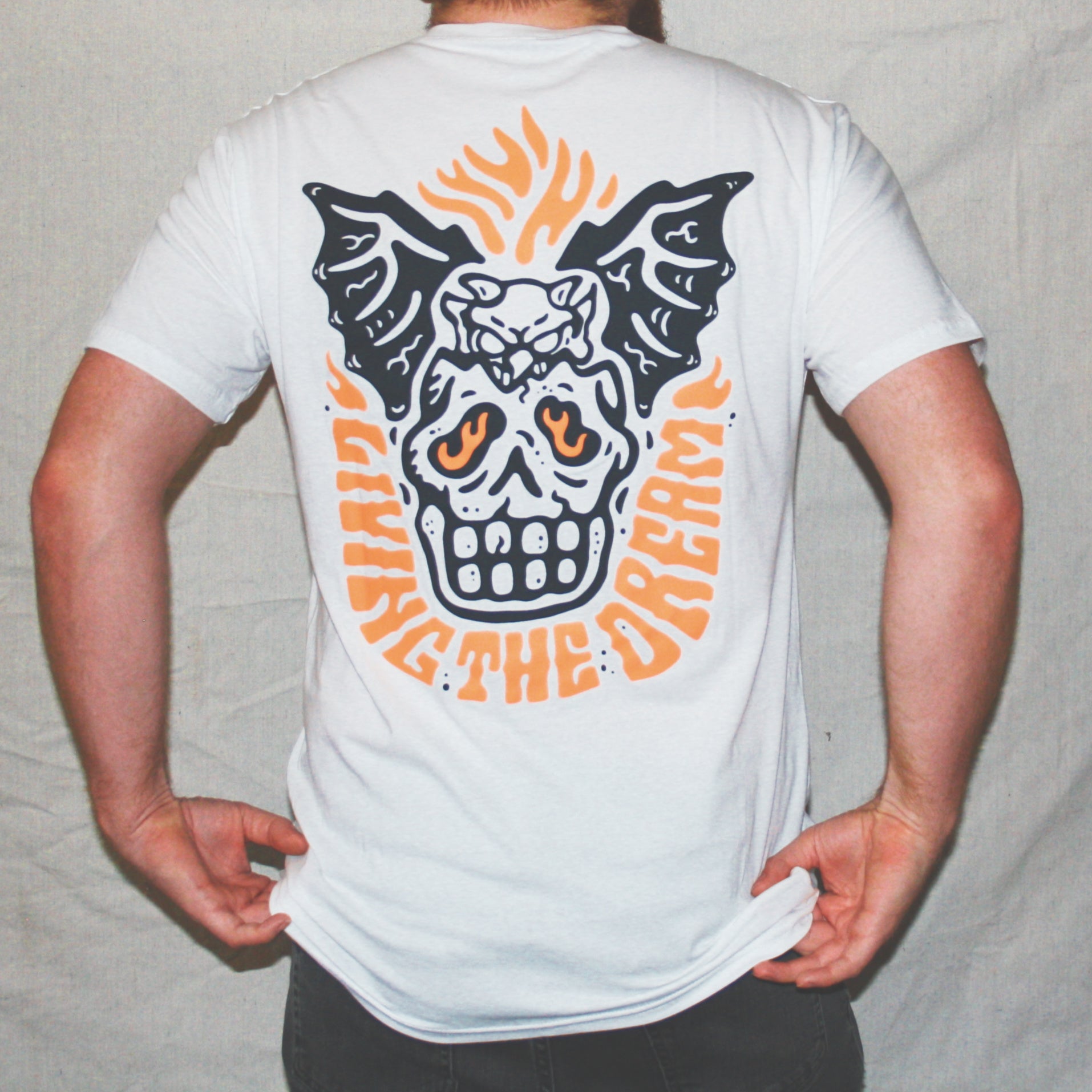 White graphic t-shirt made in Canada from a soft 100% ring-spun cotton, featuring a bold hand-drawn design of a broken skull with a winged demon peaking out the top, screen printed in black and bright orange ink. Perfect for those who appreciate lowbrow, tattoo-style artwork.