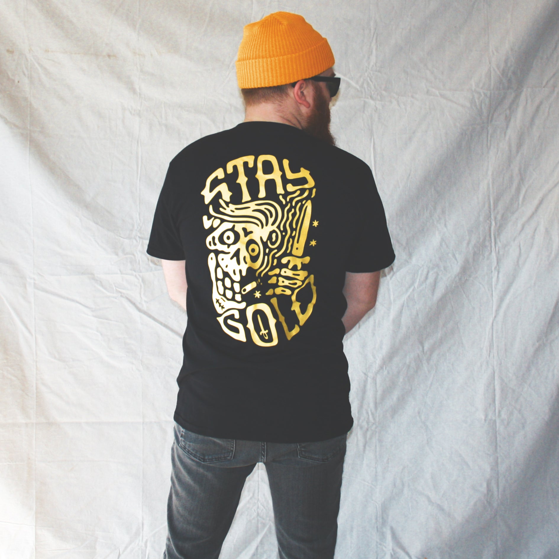 Black graphic t-shirt made in Canada from a soft 100% ring-spun cotton, featuring a bold hand-drawn design of a smiling skeleton holding a switchblade with the words “stay gold,” screen printed in yellow ink. Perfect for those who appreciate lowbrow, edgy artwork.