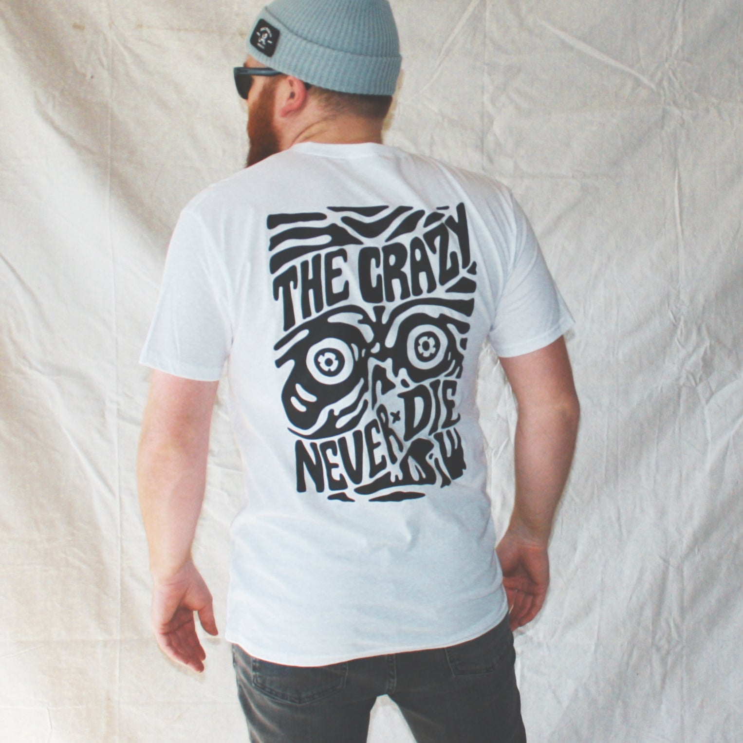 White graphic t-shirt made in Canada from a soft 100% ring-spun cotton, featuring a bold hand-drawn design of a zombie face surrounded by the words “the crazy never die,” screen printed in black ink. Perfect for those who appreciate lowbrow, horror-inspired artwork.